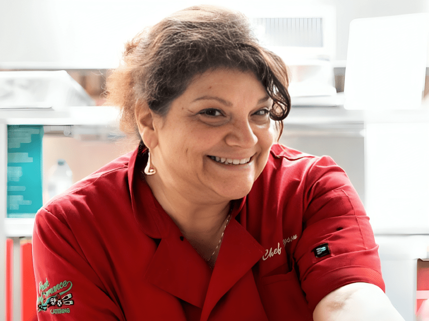 A woman wearing a red chef coat smiles while standing in a kitchen.