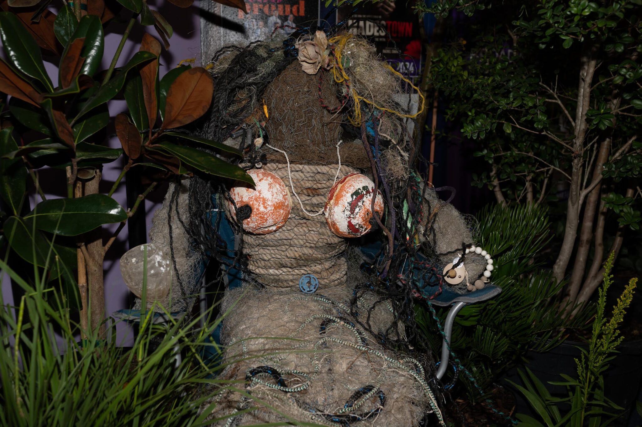 A life-sized figure made from rope, netting, and debris, with orange buoys for breasts, is seated among plants and greenery.