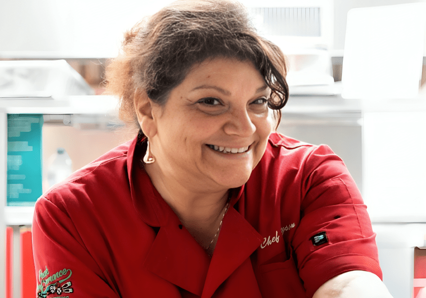 A woman wearing a red chef coat smiles while standing in a kitchen.