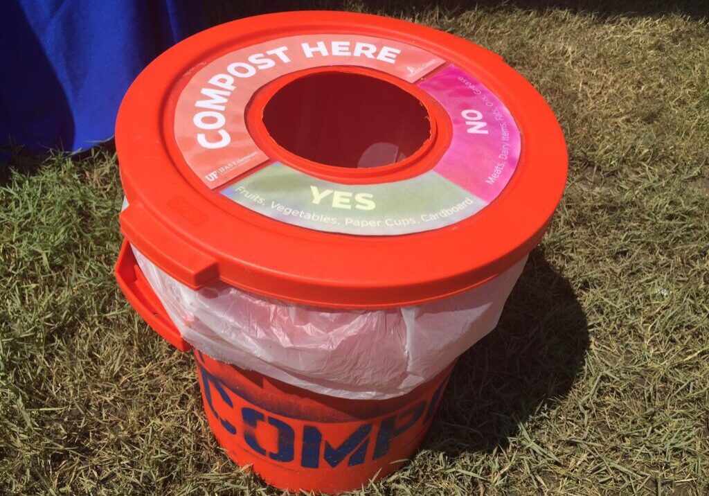 Red compost bin with a labeled lid indicating "YES" for fruits, vegetables, and paper cups, and "NO" for meat, dairy, and plastic. The bin is placed on grass outdoors.