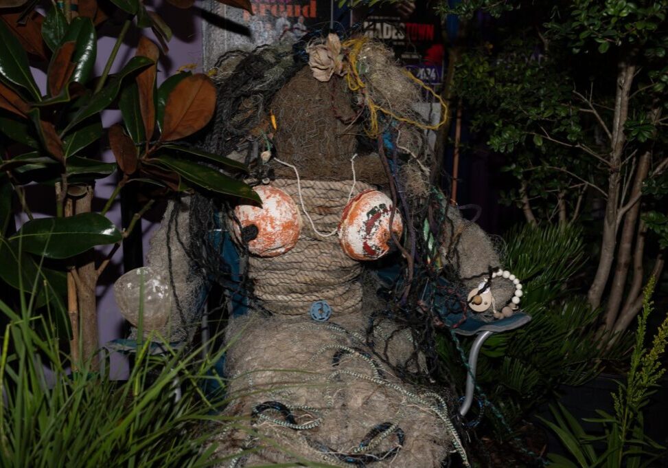 A life-sized figure made from rope, netting, and debris, with orange buoys for breasts, is seated among plants and greenery.